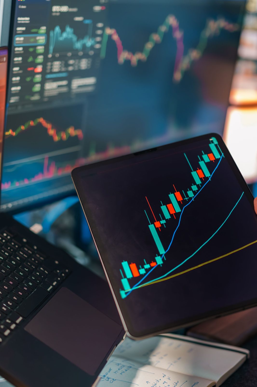 Rising candlestick chart on a tablet in front of live dashboard screens